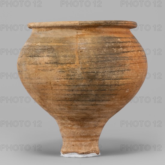 Beaker, unknown Northern Mesopotamian workshop, stemmed beakers; clay, bulging bowl with an upturned flat narrow rim, set on a low cylindrical stem terminating in a small damaged foot. Horizontal traces of polishing on the surface. 1st half of the 3rd millennium BC; Early Jazira III period ca. 2600–ca. 2350 BC. Tell Jassa el Gharbi North-Eastern Syria, Northern Mesopotamia Khabur Basin. Vessel; height 11.5 cm, width 11.6 cm, weight 264 g., A rounded earthenware vessel with a narrow foot and flared rim sits against a neutral gray background; the pot shows a weathered surface with horizontal striations and patches of varied tones including tan, brown, ochre, and darker grayish-brown areas, with a few lighter worn spots and a small band of white material at the base.