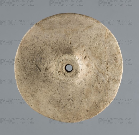 Wheel from a model wagon, unknown northern Mesopotamian workshop, terracotta model wheel with a full disc of lenticular cross-section and a protruding modelled hub on both sides for the axle, c. 2600–c. 2350 BC Early Jazira III / Early Dynastic III period, Northern Mesopotamia Khabur basin, Tell Jassa el Gharbi, Syria, model/toy, height 10 cm, width 3.74 cm, weight 114 g., A roughly circular, flat disc-shaped object with a central round hole, showing a textured surface with irregular marks and small pits; the disc is a beige to light tan color with areas of darker brownish-gray speckling and slight discoloration, set against a smooth medium gray background.
