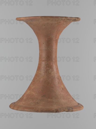 Vessel base. Funnel-shaped vessel base, narrowest at about 2/3 of the height. Both rims are oval in cross-section, slightly upturned. The upper rim has a slightly wavy line. Made of Nile clay. Outer surface and surface of the upper rim are smoothed, covered with a layer of engobe and polished; inner surface smoothed. Vessel color: red. Vessel glued together from several fragments; gaps filled with plaster. Surface eroded in places. Comparative material: ca. 2686–ca. 2181 BC; Old Kingdom, Edfu; vessel, height 23.9 cm, diameter 16.9 cm rim, diameter 19.6 cm bottom., A single terracotta object shaped like an hourglass with a flared rim and base, showing a rough, mottled surface in shades of reddish-brown and muted orange with patches of lighter beige and darker brown, small pits and irregularities across its body, a gently curved narrow middle section expanding into wide, rounded edges, and a neutral gray background.
