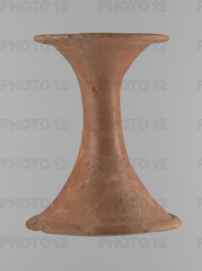 Vessel base. Funnel-shaped vessel base, narrowest at about 2/3 of the height. Both rims are oval in cross-section, slightly upturned. The upper rim has a slightly wavy line. Made of Nile clay. The outer surface and the surface of the upper rim are smoothed, covered with a layer of engobe and polished; the remaining inner surface is smoothed. Vessel color: red. Vessel glued together from several fragments; gaps filled with plaster. The surface is eroded in places. Comparative material: Reisner, A history of Giza necropolis, vol. I, London 1942, fig. 282, C,4240, ca. 2686–2181 BC; Old Kingdom, Edfu, vessel, height 23.9 cm, diameter 16.9 cm rim, diameter 19.6 cm bottom., A single hourglass-shaped clay object centered against a light gray background, with a warm reddish-brown surface showing patches of darker brown and small areas of pale beige wear; the top and bottom rims flare outward and are slightly irregular, and the midsection narrows smoothly with a textured, pitted finish and subt...