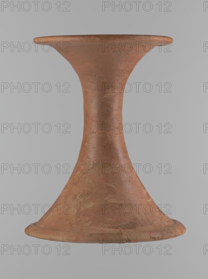 Vessel base. Funnel-shaped vessel base, narrowest at about 2/3 of the height. Both rims are oval in cross-section, slightly upturned. The upper rim has a slightly wavy line. Made of Nile clay. The outer surface and the surface of the upper rim are smoothed, covered with a layer of engobe and polished. The remaining inner surface is smoothed. Vessel color: red. Vessel glued together from several fragments; gaps filled with plaster. The surface is eroded in places. Comparative material: Reisner, A history of Giza necropolis, vol. I, fig. 282, C,4240, ca. 2686–ca. 2181 BC; Old Kingdom, Edfu, vessel. Height 23.9 cm; diameter 16.9 cm rim; diameter 19.6 cm bottom., A single hourglass-shaped terracotta object with a flared top and base, showing a textured surface with irregular patches and spots; colors visible include shades of reddish-brown, earthy orange, and small areas of darker brown, set against a neutral light gray background.