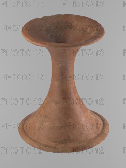 Vessel base, Funnel-shaped vessel base, narrowest at about 2/3 of the height. Both rims are oval in cross-section, slightly upturned. The upper rim has a slightly wavy line. The vessel is made of Nile clay. The outer surface and the surface of the upper rim are smoothed, covered with a layer of engobe and polished. Only the remaining inner surface is smoothed. Vessel color - red. Vessel glued together from several fragments. Gaps filled with plaster. The surface of the vessel is eroded in places. Comparative material: ca. 2686–ca. 2181 BC; Old Kingdom, Edfu, vessel, height 23.9 cm, diameter 16.9 cm rim, diameter 19.6 cm bottom, An object shaped like a tall hourglass with a flared rim and a wide base, made of a coarse, earthy material showing rough and smooth patches, in shades of reddish-brown and terracotta with darker brown and tan areas, sitting against a plain light gray background; the surface has irregular textures, small pits, and worn spots, and the rim curves outward while the...