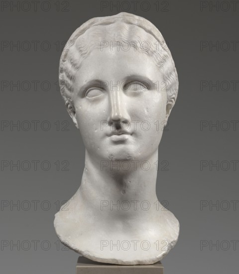 Head of a woman Berenice II or Arsinoe II, unknown creator; Greek sculpture, female portrait, Hellenistic period, 330–30 BC; Alexandria; sculpture; restitution Przeworsk, A white marble bust of a woman's head and neck is shown against a neutral gray background; the sculpture features wavy hair pulled back with a headband, smooth facial features with subtle relief around the eyes and mouth, and a gently rounded chin and long neck, with the lower edge of the bust rough and uneven resting on a small rectangular base; visible colors include white, off-white, gray, and beige.
