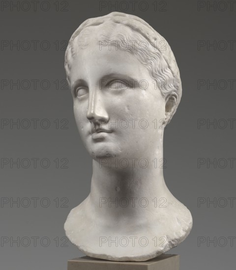 Head of a woman Berenice II or Arsinoe II, Greek Hellenistic sculpture, female portrait, 330–30 BC Hellenistic period, Alexandria; sculpture; restitution Przeworsk, A smooth white marble bust of a head and neck displayed against a neutral gray background, showing stylized wavy hair pulled back and a simple headband, subtly carved facial features with closed or blank eyes, a straight nose, softly rounded cheeks, and a small, closed mouth, with visible surface texture and slight imperfections on the marble, a tapered neck leading to a rough-edged base, and overall colors of white and shades of gray.
