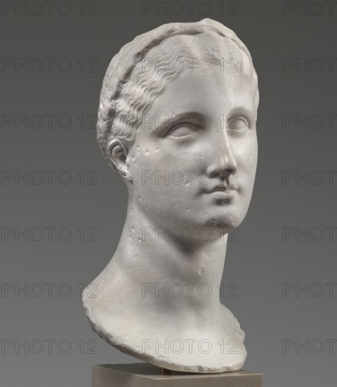 Head of a woman Berenice II or Arsinoe II, unknown author, Greek sculpture, female portrait, 330–30 BC; Hellenistic period, Alexandria, sculpture, A carved pale stone bust of a head and neck set against a smooth gray background, showing a stylized hairline with wavy carved strands and a headband, smooth facial features with closed or blank eyes, a straight nose, and a small closed mouth, with subtle surface pitting and weathering visible on the cheek and neck, mounted on a small square base; visible colors include off-white and light gray on the sculpture and medium gray in the background.