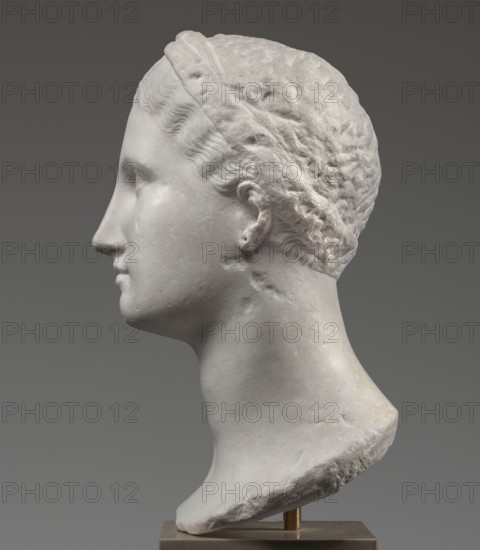 Head of a woman Berenice II or Arsinoe II, Scen. Gal. Class., restitution Przeworsk; Greek sculpture, female portrait, 330–30 BC; Hellenistic period; Alexandria; sculpture, A marble sculpted head in profile viewed from the left, showing a smooth pale stone face and neck with carved wavy hair held by a textured headband; the ear and jawline are visible and the chin projects slightly, and the hair surface shows carved ridges and rougher stone texture toward the back; the sculpture rests on a small base and the background is a solid gray. Colors visible include white, off-white, pale gray, darker gray, and muted brown on the base.