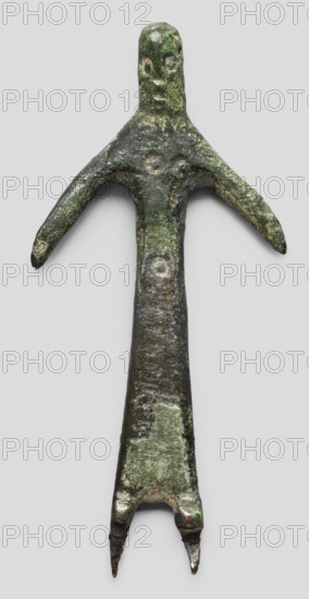 Votive figurine of a woman, votive offering, bronze, female figurine, 1st half of the 6th century BC, Etruria; height 6.1 cm, width 2.9 cm, depth 0.5 cm., A weathered green and dark brown metal figure resembling a simplified human shape with an elongated body, a rounded head with shallow eye and mouth impressions, a pair of outstretched arms, and a tapered lower body ending in two pointed prongs; the surface shows rough texture, patches of lighter green corrosion and darker brown areas, and several shallow circular indentations on the torso and head.