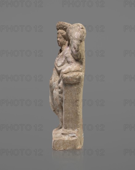 Terracotta figurine of Eros with a goose. Terracotta. Eros mythology, Greek mythology, figurines from a mold. Figurine of a standing Eros. The figure, positioned frontally, slightly leaning back and facing left, stands on a low rectangular base. On the left he leans against a small column, and on the right he holds a goose standing nearby by the neck. The left hand, clasped at the elbow, rests on the top of the column, holding a short cloak halfway up. The cloak, fastened with a fibula over the right shoulder, covers the chest and forms folds and flows along the small column. The hair, arranged in curls, is covered in the upper part by a high diadem. The outstretched wings, with straight, defined feathers, are slightly tilted back. Scratches faded, traces of polychrome. 2nd half of the 1st century BC. Asia Minor place of origin. Figurine. Height 14 cm; width 10.2 cm; depth 3.4 cm; weight 0.18 kg., A weathered carved figure in muted brown and beige tones stands against a gray background...