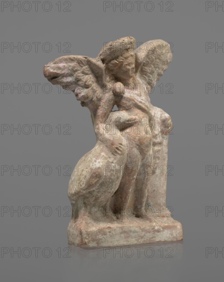 Terracotta figurine of Eros with a goose. Figurine of a standing Eros, positioned frontally, slightly leaning back and facing left, standing on a low rectangular base. He leans on a small column at the left and holds a goose by the neck at the right. The left hand, clasped at the elbow, rests on the top of the column and holds a short cloak halfway up. The cloak, fastened with a fibula over the right shoulder, covers the chest and forms folds and flows along the column. Hair arranged in curls and covered in the upper part by a high diadem. Outstretched wings with straight, defined feathers slightly tilted back. Scratches faded, traces of polychrome. Terracotta; figurine from a mold. 2nd half of the 1st century BC, Asia Minor. Height 14 cm, width 10.2 cm, depth 3.4 cm, weight 0.18 kg., A weathered stone figurine with a rough surface showing tones of beige, pale pink, muted green, and gray stands on a rectangular base; the figure has a rounded head with indistinct facial features, a pair...