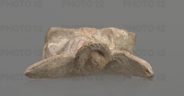 Terracotta figurine of Eros with a goose. Figurine of a standing Eros: the figure, positioned frontally, slightly leaning back and facing left, stands on a low rectangular base. On the left he leans against a small column; on the right he holds a goose by the neck. The left hand, clasped at the elbow, rests on top of the column holding a short cloak halfway up. The cloak, fastened with a fibula over the right shoulder, covers the chest and forms folds and flows along the small column. Hair arranged in curls, covered in the upper part by a high diadem. Outstretched wings with straight, defined feathers are slightly tilted back. Scratches faded; traces of polychromy. Terracotta, from Asia Minor; 2nd half of the 1st century BC. Figurine from a mold. Height 14 cm, width 10.2 cm, depth 3.4 cm, weight 0.18 kg., A weathered stone or clay object rests on a smooth gray surface against a neutral gray background, showing a rough, textured surface with irregular shapes and worn edges; the object h...