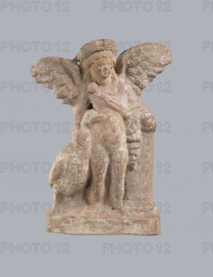 Terracotta figurine of Eros with a goose. Terracotta, figurine from a mold, 2nd half of the 1st century BC, Asia Minor. Figurine of a standing Eros positioned frontally, slightly leaning back and facing left, standing on a low rectangular base. He leans on a small column at his left and holds a goose by the neck at his right. The left hand, clasped at the elbow, rests on the top of the column and holds a short cloak halfway up; the cloak, fastened with a fibula over the right shoulder, covers the chest and forms folds along the column. Hair arranged in curls, covered in the upper part by a high diadem. Outstretched wings with straight, defined feathers, slightly tilted back. Scratches faded, traces of polychrome. Height 14 cm, width 10.2 cm, depth 3.4 cm, weight 0.18 kg., A small weathered sculpture of a seated winged figure on a rectangular base, rendered in earthy tones of beige, brown, pale pink, and hints of gray; the figure has a rounded head with a headdress, textured wings exten...