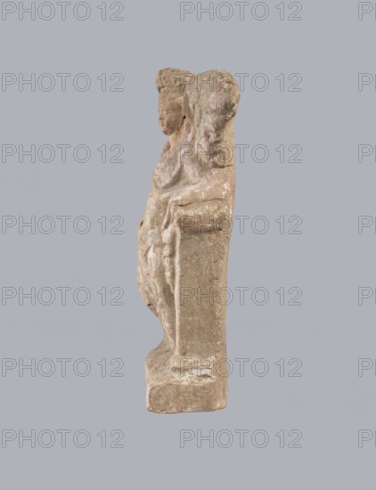 Terracotta figurine of Eros with a goose. Terracotta; figurine of a standing Eros produced from a mold. The figure stands frontally, slightly leaning back and facing left, on a low rectangular base. On the left he leans against a small column; on the right he holds a nearby goose by the neck. The left hand, clasped at the elbow, rests on the top of the column and holds a short cloak pulled halfway up. The cloak, fastened with a fibula over the right shoulder, covers the chest and forms folds that flow along the column. Hair arranged in curls, covered in the upper part by a high diadem. Outstretched wings with straight, defined feathers, slightly tilted back. Scratches faded; traces of polychromy. 2nd half of the 1st century BC, Asia Minor. Height 14 cm; width 10.2 cm; depth 3.4 cm; weight 0.18 kg., A weathered stone sculpture shown from the side depicting a figure with a rounded head and textured hair leaning against a vertical slab, with visible carved folds suggesting draped clothing...