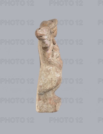 Terracotta figurine of Eros with a goose. Terracotta, figurine from a mold, figurine of a standing Eros. The figure, positioned frontally, slightly leaning back and facing left, stands on a low rectangular base. On the left he leans against a small column; on the right he holds a goose by the neck. The left hand, clasped at the elbow, rests on the top of the column, holding a short cloak halfway up. The cloak, fastened with a fibula over the right shoulder, covers the chest and forms folds and flows along the small column. Hair arranged in curls, covered in the upper part by a high diadem. Outstretched wings with straight, defined feathers, slightly tilted back. Scratches faded, traces of polychrome. 2nd half of the 1st century BC, Asia Minor; height 14 cm, width 10.2 cm, depth 3.4 cm; weight 0.18 kg., A weathered sculpture of an animal-like figure shown in profile against a plain gray background, with a rounded snout, curved ears, and a pronounced neck and back; the surface appears ro...