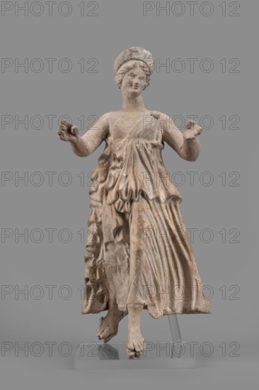 Figurine of the goddess Nike;, Nike mythology, Greek deity; female terracotta figurine; 1st half of the 2nd century BC; found at Myrina; height 27 cm, width 12 cm, depth 11 cm, weight 0.47 kg., A weathered beige stone statue of a standing female figure wearing a flowing, draped garment with detailed folds, a shawl-like piece hanging at one side, and a rounded headpiece; the figure has outstretched arms with hands partially open, a calm facial expression, and braided hair; the statue stands on a clear display support and base against a uniform gray background, with visible surface texture and subtle darker and lighter beige variations.