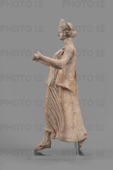 Figurine of the goddess Nike, Nike mythology, Greek deity, female figurine, 1st half of the 2nd century BC, Myrina site of finding, figurine, height 27 cm, width 12 cm, depth 11 cm, weight 0.47 kg, A weathered statue of a robed female figure shown in profile, made of a light beige to pale brown stone with areas of darker brown patina and faint traces of reddish tones, standing on a gray rectangular base with a metal support visible; she faces left with her hair pulled back under a cap, wearing a draped garment that shows carved folds and pleats cascading down the front and side, arms extended forward with hands clasped together, bare feet stepping forward with visible toes, and the surface shows rough texture and small abrasions.