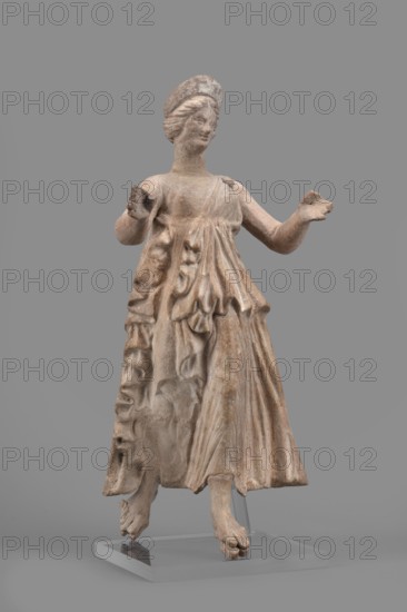 Figurine of the goddess Nike;, repossession Silesia; Nike mythology; Greek deities; female figurine; 1st half of the 2nd century BC; Myrina place of finding; terracotta figurine; height 27 cm, width 12 cm, depth 11 cm, weight 0.47 kg, A weathered statue of a standing figure wearing a long draped garment with detailed folds and ruffles, shown atop a clear rectangular base; the figure has both arms raised with open hands, visible feet beneath the hem, and a rounded headpiece. The surface shows tones of beige, tan, light brown, and hints of gray against a mid-gray background.