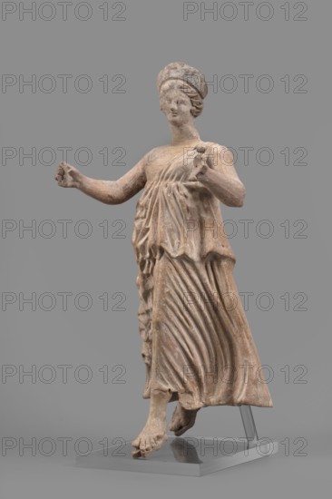 Figurine of the goddess Nike, repossession Silesia, Nike mythology, Greek deity, female figurine, 1st half of the 2nd century BC, Myrina site of finding, terracotta figurine, height 27 cm, width 12 cm, depth 11 cm, weight 0.47 kg, A weathered terracotta statue of a standing female figure on a clear rectangular base, shown against a solid gray background, with a few areas of faded reddish-brown and beige surface tones; she wears a draped, flowing dress with pronounced vertical and diagonal folds and a short overgarment gathered at the waist, a rounded headpiece, and her hair styled in waves; her arms are extended outward with hands partially closed, one foot is slightly raised while the other rests on the base, and the sculpture shows surface texture variations and small chips.