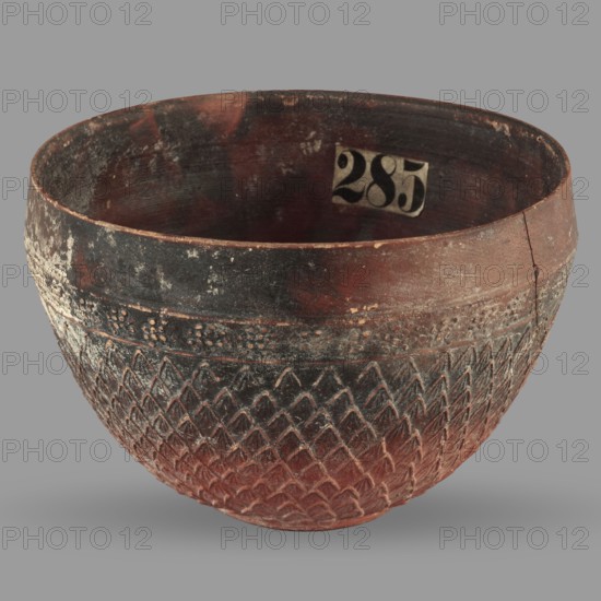 Megarian bowl, unknown Ionian workshop, classified Gallic scenography, end of 2nd–beginning of 1st century BC, Ionia Turkey/Greece, vessel; height 8.5 cm, width 13.1 cm, weight 0.178 kg., A rounded ceramic bowl with a textured exterior showing a repeating diamond pattern and a band of small raised dots near the rim, exhibiting worn surfaces and chips. The bowl displays a mix of red, dark brown, black, and traces of lighter beige on its exterior and interior. The interior appears darker with areas of mottled discoloration, and the rim shows signs of wear and a visible crack on one side. The background is a uniform gray.