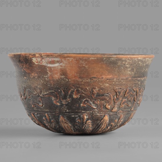 Megarian bowl, KIPBEI workshop; 2nd half of the 2nd century BC; possibly Smyrna; vessel; weight 0.120 kg., A shallow, rounded ceramic bowl with a flared rim, showing worn and textured surfaces in shades of brown, black, and reddish-orange; the upper portion is lighter brown with areas of darker discoloration, while the lower portion is predominantly blackened with reddish-orange highlights where carved decorations appear; a band of raised, stylized floral and foliate motifs wraps around the middle, and a row of carved leaf-like forms adorns the base; the bowl sits on a smooth gray surface against a plain gray background.