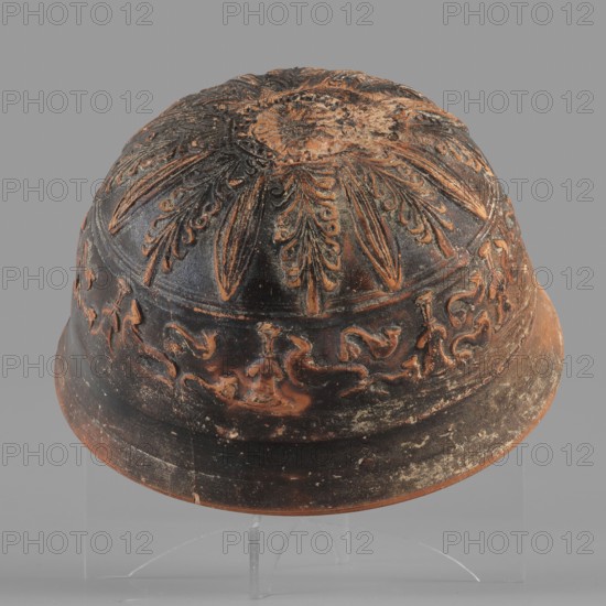 Megarian bowl, KIPBEI workshop, 2nd half of the 2nd century BC, Smyrna, vessel, Megarian bowl, weight 0.120 kg, A domed ceramic object sits against a plain gray backdrop, showing a textured surface with ornate raised decorations; the top has a radial floral or sunburst pattern with elongated petal-like elements, and the midband features a continuous frieze of flowing, scroll-like and stylized animal or bird figures in relief. The object displays a worn, earthy color palette with dominant dark brown to black areas and contrasting terracotta and rusty orange highlights where the raised decoration and edges show through; there are patches of lighter beige and pale wear on the surface. The base edge is slightly flared and shows surface abrasion and small chips, and the piece rests on a clear support that is partially visible beneath it.