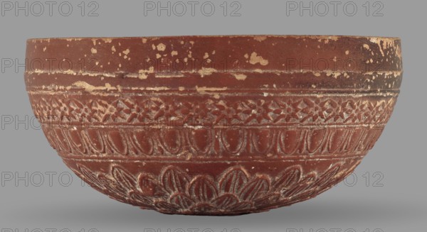 Megarian bowl, Monogramist's Workshop, 2nd half of the 2nd century BC, Ephesus discovered in, Ionia place of origin, vessel, weight 0.134 kg, A shallow rounded bowl photographed against a plain gray background, with a worn reddish-brown surface showing patches where a lighter tan color has flaked away; the bowl features several horizontal bands of carved decorative patterns including a band of small geometric motifs, a band of repeated petal or teardrop shapes, and a lower band with larger overlapping petal motifs, all highlighted by the lighter tan showing in the recessed carving.