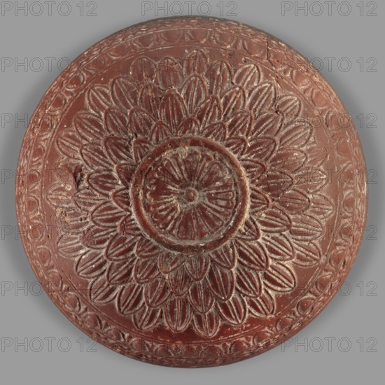 Megarian bowl, Monogramist's Workshop, 2nd half of the 2nd century BC, Ephesus, Ionia; vessel; weight 0.134 kg, A circular carved object with a raised floral medallion design occupying the center, showing layered petal shapes radiating outward and a round central rosette, all rendered in a reddish-brown color with lighter worn highlights revealing a paler tone, set against a neutral gray background; the surface shows carved linear and scalloped border patterns around the rim and areas of surface wear and small chips.