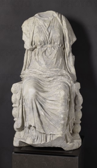 Statue of the goddess Cybele on a throne, Cybele myth., statue of a deity, Roman sculpture, 1st century BC–1st century AD, Asia Minor, sculpture; height 84 cm, width 43 cm, depth 39 cm, weight 181 kg, A headless stone statue of a seated draped figure sits on a blocky base against a dark background, showing folded and gathered garments with detailed carved folds and flowing drapery in pale grey and off-white tones, with areas of subtle discoloration and weathering; the figure's arms are partially present beneath the cloth, and the seat has rounded carved elements visible at the sides, while the base beneath the statue is a darker stone with a smooth surface in charcoal and deep grey colors.
