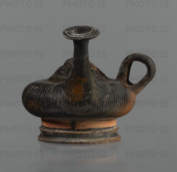 Black-finned guttus, repossession Silesia, guttus, Greek pottery, black-finned pottery. Guttus with a high foot composed of a profiled lower part and a cylindrical, narrower upper part this part is not covered with the finish. Body shaped like a flattened sphere with vertical grooves. A band-shaped handle on the right side of the vessel. Long neck with a wide, flared rim. On the upper surface is a convex medallion, surrounded by a single band, depicting the head of Heracles in a lion's skin. Round face, massive nose, wide mouth, large almond-shaped eyes. The lion-skin and mane frame Heracles' face. The finish ranges from black to shiny brown. Condition: the handle and neck are cracked and glued; cracks and abrasions in the finish. 3rd century BC, Calvi place of manufacture, vessel, guttus, height 10.7 cm, diameter 11.5 cm., An ancient-looking ceramic vessel with a rounded body, a tall narrow neck flaring into a small open mouth, and a single looped handle; the surface shows ribbed text...