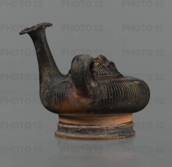 Black-finned guttus, guttus with a high foot composed of a profiled lower part and a cylindrical, narrower upper part this part is not covered with the finis. The body is shaped like a flattened sphere with vertical grooves. A band-shaped handle is located on the right side of the vessel. Long neck with a wide, flared rim. On the upper surface is a convex medallion, surrounded by a single band, depicting the head of Heracles in a lion's skin. Round face, massive nose, wide mouth, large almond-shaped eyes. The skin and mane frame Heracles' face. The finis ranges from black to shiny brown. Condition: the handle and neck are cracked and glued; cracks and abrasions in the finial. 3rd century BC, Calvi, vessel, guttus, height 10.7 cm, diameter 11.5 cm, An ancient-looking ceramic vessel sits against a plain gray background; it has a rounded, ribbed body with a flattened base and a narrow raised foot, showing earthy terracotta and dark blackened surfaces with worn areas where the terracotta s...