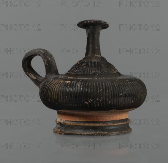 Black-finned guttus, guttus, Greek pottery, black-finned pottery. Guttus with a high foot composed of a profiled lower part and a cylindrical, narrower upper part this part is not covered with the finis. Body shaped like a flattened sphere with vertical grooves. A band-shaped handle is located on the right side of the vessel. Long neck with a wide, flared rim. On the upper surface is a convex medallion, surrounded by a single band, depicting the head of Heracles in a lion's skin. Round face, massive nose, wide mouth, large almond-shaped eyes. The lion skin and mane frame Heracles' face. The finis ranges from black to shiny brown. Condition: the handle and neck are cracked and glued; cracks and abrasions in the finial. 3rd century BC, Calvi place of manufacture, vessel, guttus, height 10.7 cm, diameter 11.5 cm, A single rounded vessel with a short neck and a narrow flared rim sits against a plain background, showing a curved handle on one side and a ridged, bulbous body; the surface is ...