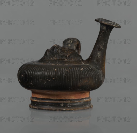 Black-finned guttus, guttus, Greek pottery, black-finned pottery. Guttus with a high foot composed of a profiled lower part and a cylindrical, narrower upper part this part is not covered with the finis. Body in the shape of a flattened sphere with vertical grooves. A band-shaped handle on the right side of the vessel. Long neck with a wide, flared rim. On the upper surface is a convex medallion, surrounded by a single band, depicting the head of Heracles in a lion's skin. Round face, massive nose, wide mouth, large almond-shaped eyes. The lion skin and mane frame Heracles' face. Finish ranges from black to shiny brown. Condition: the handle and neck are cracked and glued; cracks and abrasions in the finish. 3rd century BC, Calvi place of manufacture, vessel, guttus; height 10.7 cm, diameter 11.5 cm., A small sculpted vessel with a bulbous, ribbed body and a short flared base, shown in profile against a plain grey background; the vessel is predominantly dark brown to black with worn ar...