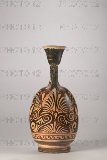 Red-figure aryballus lekythos with a depiction of Eros and a woman, Darius Painter active 340–320 BC, 340–320 BC, Apulia; vessel lekythos, aryballus lekythos, height 23.3 cm, width 10.7 cm, A single narrow-necked ceramic vessel with a flared rim sits upright on a neutral background; the vessel has a bulbous body, a slender vertical neck, and a stepped circular base. The surface shows decorative painted patterns in dark black against an orange-brown clay color, with areas of lighter beige where the paint has worn; the design includes fan-shaped motifs and curling spiral elements wrapping around the body and a band of repeating wave-like shapes near the base. The rim and upper neck have a darker black coating with small areas of flaking that reveal the lighter clay beneath, and the overall color palette includes black, orange-brown, beige, and small touches of darker brown on the base.