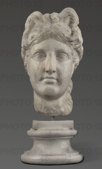 Head of the goddess Aphrodite; unknown creator; exchange with the GDR; Greek sculpture, female portrait; hair tied in a bow at the front, in a bun at the back; 332–30 BC Hellenistic period, Greece; sculpture; weight 9.74 kg, A marble sculpture of a head displayed on a pedestal, showing a face with closed or blank eyes, a straight nose, and neutral lips; the hair is carved in wavy, voluminous locks arranged atop and around the head, with some textured detailing at the sides and back. The stone surface shows subtle variations in tone and some weathered or rougher patches, especially near the chin and neck area. The pedestal and base are also marble with rounded, tiered forms. Visible colors include shades of white, off-white, light gray, and darker gray.