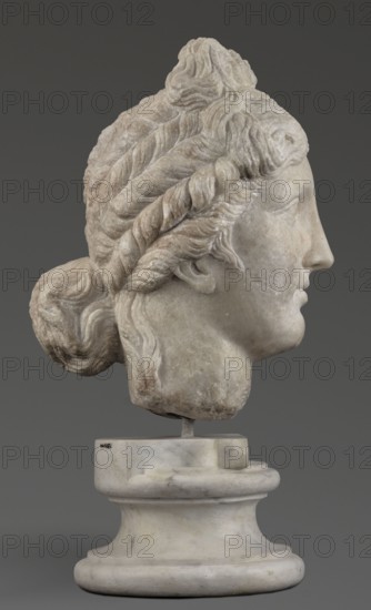 Head of the goddess Aphrodite; creator, Greek sculpture, female portrait; hair tied in a bow at the front, in a bun at the back; Hellenistic period, 332–30 BC; Greece; sculpture; weight 9.74 kg, A carved stone head of a person shown in profile mounted on a round pedestal, with detailed wavy and braided hair swept back into a bun; the sculpture has a pale off-white and light gray surface with areas of darker beige and brown discoloration, and it sits against a solid dark gray background.