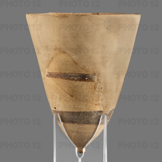 Cup, unknown Middle Assyrian workshop; Mitannian painted pottery Northern Mesopotamia, Mitanni Kingdom in Northern Mesopotamia; Eastern Gallic Scenography; 14th–13th century BC, Middle Assyrian period; vessel., A conical, beige ceramic vessel with a pointed bottom sits on a clear support against a gray background, showing a worn surface texture with scratches and small pits, a few horizontal dark brown bands and a faint darker area on one side, and subtle variations of tan and ochre tones across the body.