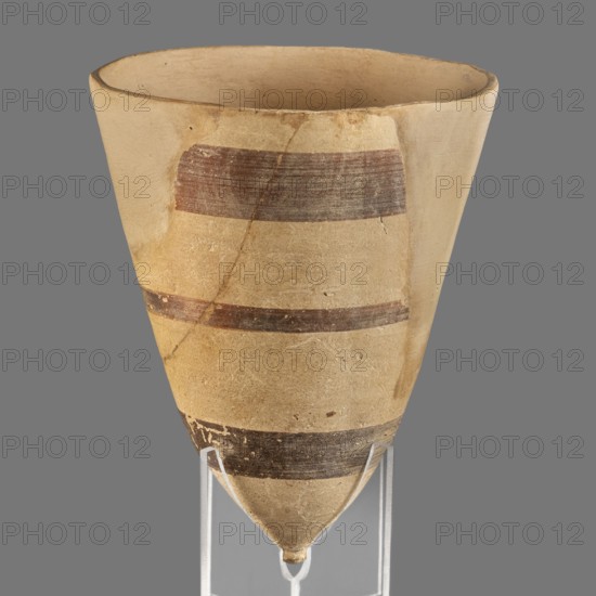 Cup, unknown Middle Assyrian workshop, Mitannian painted pottery Northern Mesopotamia, Mitanni Kingdom in Northern Mesopotamia, painted pottery, Near Eastern pottery, Eastern Gallic scenography, 14th–13th century BC; Middle Assyrian period, Northern Mesopotamia, vessel., A tall, conical ceramic vessel with a pointed base sits on a clear stand, showing a beige surface with a few horizontal bands in dark brown and reddish-brown; the rim is uneven and the body displays visible surface texture, small chips, and hairline cracks, with colors including beige, dark brown, reddish-brown, and a gray background behind the object.