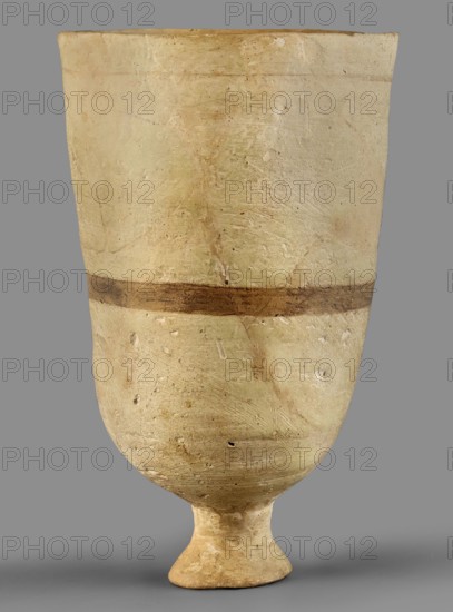 Cup, unknown Mitannian workshop; Mitannian pottery 15th–14th century BC; stemmed cup; material: pottery; Mitanni, Northern Mesopotamia Kingdom of Mitanni; Mitannian period; vessel., A tall, rounded ceramic vessel with a narrow flared foot standing against a gray background; the vessel's surface shows an uneven, textured finish in pale beige and cream tones with areas of light brown and subtle greenish tints, and a single horizontal darker brown band encircling the midsection; the surface displays small pits and scratches and varied discolorations with a slightly darker base and a lighter rim.