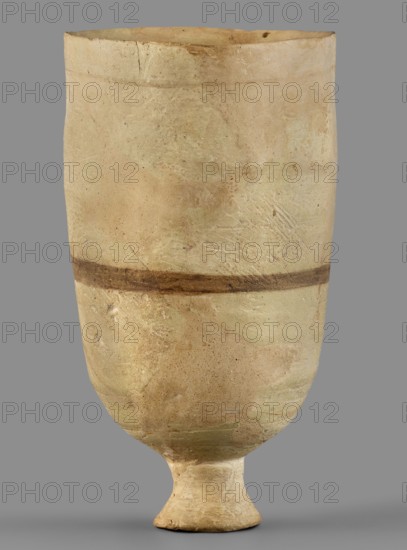 Cup, unknown Mitannian workshop, Mitannian pottery 15th–14th century BC, stemmed cup, Mitanni, Northern Mesopotamia Kingdom of Mitanni, material: pottery, function: vessel, period: Mitannian 15th–14th century BC, An upright ceramic vessel with a rounded body that tapers to a short pedestal foot, showing a matte, weathered surface in shades of beige, cream, light tan, and pale green, with a darker brown horizontal band around the midsection and scattered small darker specks and patches; the rim is slightly irregular with minor chips and the surface displays subtle texture variations, scratches, and areas of wear.