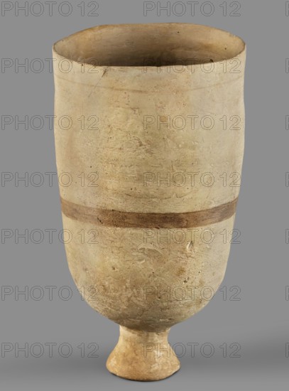 Cup, unknown Mitannian workshop, Mitannian pottery 15th–14th century BC, stemmed cup, material culture, Mitanni, Near Eastern pottery, Eastern Gallic scene, 15th–14th century BC; Mitannian period, Northern Mesopotamia Kingdom of Mitanni, vessel, A tall, cylindrical ceramic vessel with a rounded base that tapers to a short pedestal foot, shown against a plain gray background; the vessel surface is textured and worn with an overall pale beige or sandy color, a slightly darker band of brown encircles the midsection, and there are subtle variations in tone including light tan and off-white patches as well as small areas of darker brown and faint greenish discoloration.