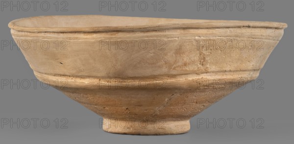Bowl, unknown Middle Assyrian workshop; Middle Assyrian pottery, bowls on a ring-shaped foot; bowl with broken profile; vertical wall above the break slightly inclined and decorated with two grooves; ring-shaped foot; 13th–12th century BC; Middle Assyrian period, Northern Mesopotamia; vessel., A shallow, wide bowl with a rounded rim and a small circular foot, shown against a plain background; the bowl surface has a textured, slightly rough appearance with visible tool marks and irregularities, and a raised band running around its midsection; colors visible include beige, tan, light brown, and hints of darker brown in weathered areas.
