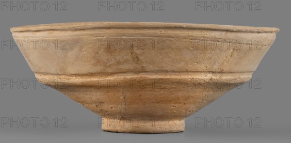 Bowl, unknown Middle Assyrian workshop; Middle Assyrian pottery; bowls on a ring-shaped foot; Near Eastern pottery; Eastern Gaulish scene; with a broken profile; vertical wall above the break slightly inclined and decorated with two grooves; ring-shaped foot; 13th–12th century BC; Middle Assyrian period, Northern Mesopotamia; vessel, A shallow, wide bowl-shaped object made of textured material with an unfinished surface, shown against a plain gray background; the object has a flared rim, a slightly recessed band around its midsection, and a tapered lower portion ending in a short circular base; visible surface irregularities include small pits, scratches, and subtle tool marks; the colors present are various shades of tan, beige, light brown, and hints of ochre.