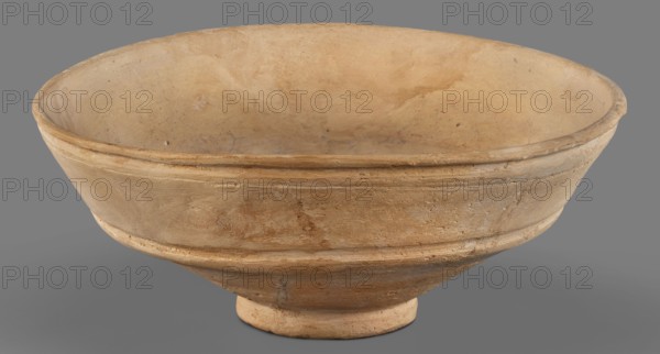 Bowl, unknown Middle Assyrian workshop; Middle Assyrian pottery; bowl on a ring-shaped foot; Near Eastern pottery; with a broken profile; vertical wall above the break slightly inclined and decorated with two grooves; ring-shaped foot; 13th–12th century BC; Middle Assyrian period, Northern Mesopotamia; vessel, A shallow, wide bowl with a rounded rim and a short circular foot, shown from the front on a neutral gray background; the bowl surface appears worn and textured with visible imperfections and small chips, and the interior and exterior show subtle variations in color including beige, tan, light brown, and hints of ochre, with some areas appearing slightly darker or stained.