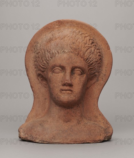 Votive head, Hellenistic period, 332–30 BC; Central Italy; sculpture; height 30 cm, width 22.5 cm, depth 17.5 cm, weight 6.1 kg., A terracotta-colored sculpted head with a rounded halo-like backplate, showing a stylized face with closed or blank eyes, a straight nose, and a small mouth, framed by textured, wavy hair; the piece rests on a roughened base and displays surface speckling and darker brown and blackish flecks against the dominant reddish-brown and tan tones, set against a plain light gray background.