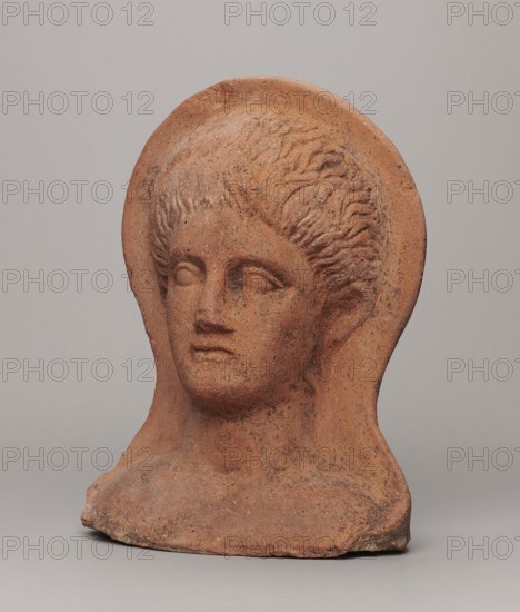Votive head, Hellenistic period, 332–30 BC, Central Italy; sculpture; height 30 cm, width 22.5 cm, depth 17.5 cm, weight 6.1 kg., A terracotta-colored sculpted head of a person with stylized hair set within a rounded halo-like backplate, showing a neutral facial expression, closed or lightly defined eyes, straight nose, and full lips; the surface has a rough, speckled texture with darker brown and black mottling, and the base shows irregular edges and wear.