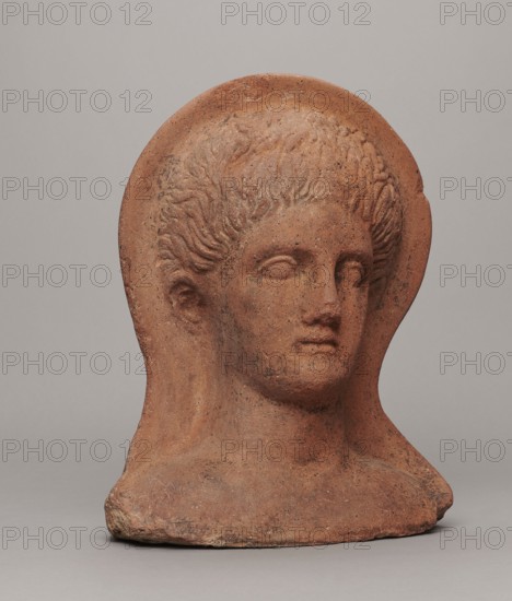 Votive head, Hellenistic period, 332–30 BC, Central Italy; sculpture; height 30 cm, width 22.5 cm, depth 17.5 cm, weight 6.1 kg., A sculpted bust of a human head and upper shoulders, made of a reddish-brown terracotta material with visible speckling and surface texture; the face has softly modeled features with closed or blank eyes, a straight nose, and a slightly closed mouth, and the hair is carved in wavy locks framing the forehead and temples; the bust sits on a broad base and shows some rough, chipped edges and darker spots; the background is a smooth, neutral light gray.
