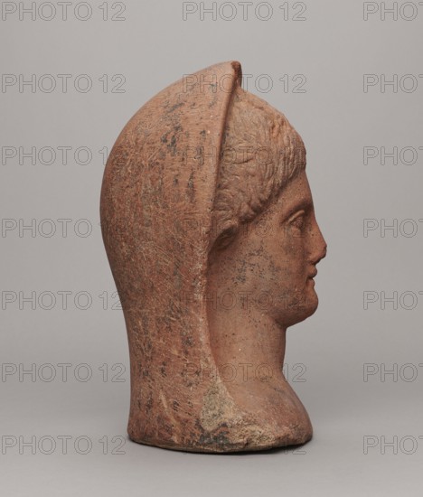 Votive head, unknown creator; Hellenistic period, 332–30 BC; Central Italy; sculpture; height 30 cm, width 22.5 cm, depth 17.5 cm, weight 6.1 kg., A terracotta-colored sculpted head in profile facing right, with a rounded helmet-like covering that extends down the back and sides, showing textured surface marks and patches of darker brown and black discoloration, a smooth forehead and stylized short hair texture, a defined nose, lips, and chin, and a similarly colored neck and base resting on a neutral pale gray surface with a pale gray background.
