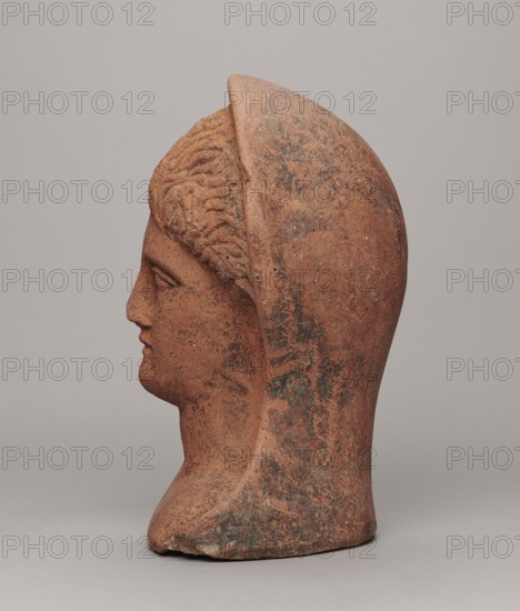 Votive head, Hellenistic period, 332–30 BC; Central Italy; sculpture; height 30 cm, width 22.5 cm, depth 17.5 cm, weight 6.1 kg; provenance: votive function., A terracotta-colored sculpted head shown in profile, with textured hair and a smooth hood-like covering extending from the forehead over the back of the head, displaying patches of darker brown and greenish discoloration and lighter worn areas, set against a uniform light gray background.