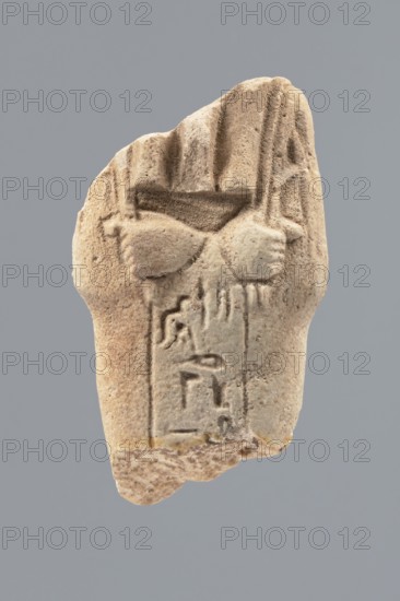 Ushabti - fragment, Mummy-shaped figurine; flat pillar on the back. Preserved central fragment from shoulder right arm damaged to hip height. Arms crossed on the chest, right folded over left; only hands visible from under the shroud. Hoes in both hands with slightly curled ends. In the right hand, a smooth rope bearing a trapezoidal basket with a semicircular handle slung over the left shoulder. Convex tools; engraved basket. Remains of a three-part smooth wig; at the back the wig is slightly wider than the pilaster. Fragmentarily preserved straight, long beard. Engraved inscription placed in a single column on the front of the legs, text bounded by two vertical lines. Excavated at Tell Atrib, shabti; height 6 cm, width 3.4 cm., A weathered beige stone fragment with a textured, grainy surface sits against a flat gray background; the fragment shows carved, rounded forms resembling a pair of clasped hands or rounded shapes near the top with vertical grooves and ridges above them, and a ...