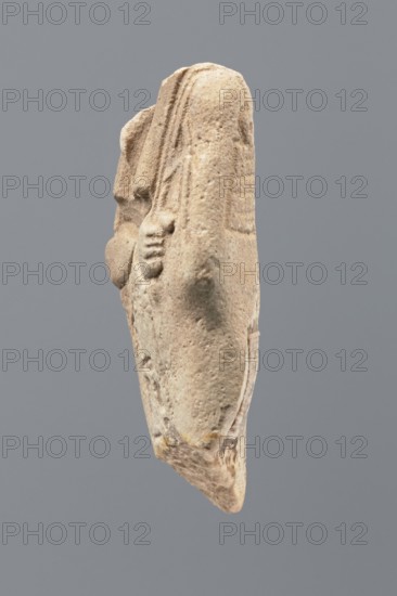 Ushabti - fragment, Mummy-shaped figurine; flat pillar on the back. Preserved central fragment from the shoulder right arm damaged to hip height. Arms crossed on the chest, right folded over left; only hands visible from under the shroud. Hoes in both hands with slightly curled ends. In the right hand a smooth rope supporting a trapezoidal basket with a semicircular handle slung over the left shoulder. Convex tools; engraved basket. Remains of a three-part smooth wig, slightly wider at the back than the pilaster. Fragmentary straight long beard. Engraved inscription placed in a single column on the front of the legs, text bounded by two vertical lines. Tell Atrib Egypt, shabti; height 6 cm, width 3.4 cm., A light beige carved stone object viewed from the side against a flat gray background, showing a rounded vertical form with textured, weathered surface and a few smooth worn areas, a partial raised relief suggesting a hand-like shape on one side, subtle linear grooves near the top, an...