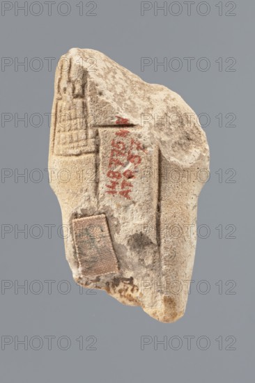 Ushabti - fragment, Mummy-shaped figurine, funerary offering. Flat pillar on the back. Preserved central fragment of the figurine—from the shoulder right arm damaged to hip height. Arms crossed on the chest, right folded over the left. Only the hands visible from under the shroud. Hoes in both hands; ends slightly curled. In the right hand a smooth rope supporting a trapezoidal basket with a semicircular handle slung over the left shoulder. Convex tools; engraved basket. Remains of a three-part smooth wig; at the back the wig is slightly wider than the pilaster. Fragmentarily preserved beard; originally straight and long. Engraved inscription placed in a single column on the front of the legs, text bounded by two vertical lines. Findspot: Tell Atrib, shabti. Dimensions: height 6 cm, width 3.4 cm., A fragment of a light beige stone with a rough, pitted surface suspended against a muted gray background, showing carved linear grooves and shallow rectangular impressions on one face, traces...